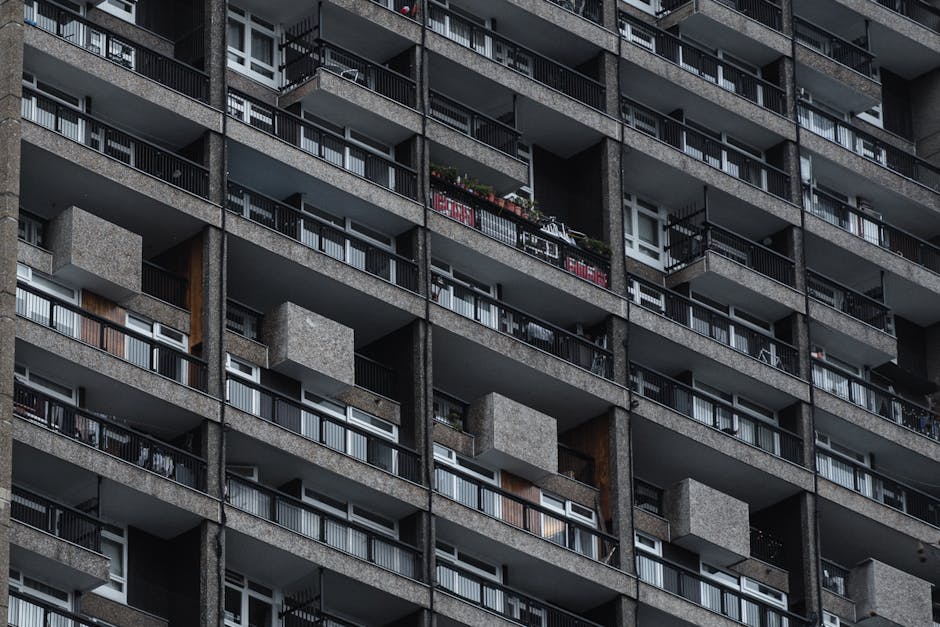 Close-up view of a multi-story residential building with numerous balconies, each with black metal railings and some with potted plants and outdoor furniture. The exterior is constructed from grey con