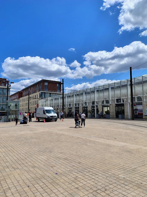 A narrow urban street scene viewed from the ground level during daytime, with a wet asphalt road marked by a central white dashed line. On both sides, there are tall, multi-storey buildings with a mix